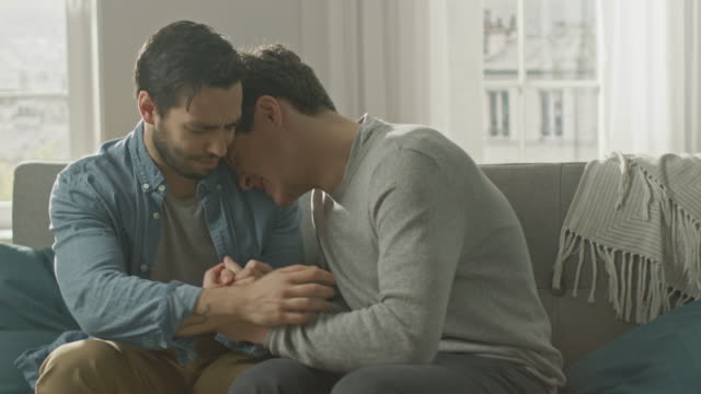 Sad-Queer-Drama-Concept.-Boyfriend-is-Unhappy-and-Depressed-About-Something.-His-Gay-Friend-is-Comforting-Him,-Holding-His-Hands.-Miserable-Man-Puts-His-Head-on-a-Shoulder-and-Cries.
