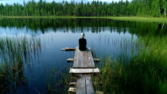 A-girl-sitting-on-the-edge-of-a-wooden-pier-near-the-smooth-water-of-a-forest-lake