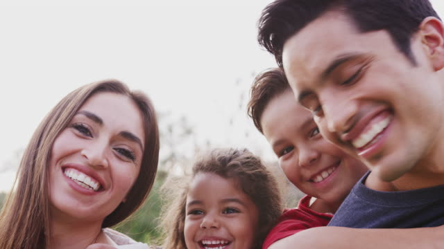 Head-and-shoulders-close-up-of-smiling-young-Hispanic-parents-piggybacking-their-children-in-park
