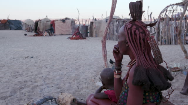 4K-side-view-of-Himba-woman-in-traditional-dress-with-young-child,-sitting-and-smoking-outside-their-hut-within-their-small-compound,-Namibia