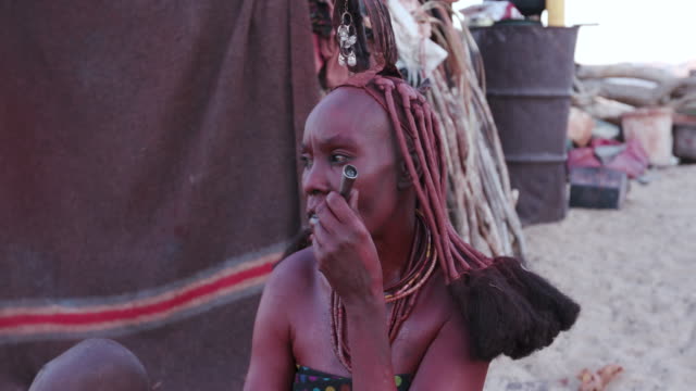 4K-Nahaufnahme-der-Himba-Frau-in-traditioneller-Kleidung-mit-Kleinkind,-rauchen-eine-Pfeife-vor-ihrer-Hütte-in-ihrer-kleinen-Anlage,-Namibia