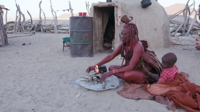 4K-Blick-auf-die-Himba-Frau-in-traditioneller-Kleidung-mit-Kleinkind,-die-einen-kleinen-Topf-auf-ein-Feuer-vor-ihrer-Hütte-in-ihrer-kleinen-Anlage,-Namibia