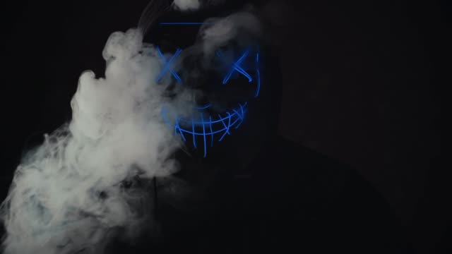 Man-with-lighting-neon-glow-mask-in-hood-and-vape-vapour-from-mask-on-black-background.-Halloween-and-horror-concept