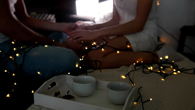 Young-and-attractive-man-and-woman-embracing-and-kissing-while-sitting-on-the-bed-in-the-dark-lit-garland-room
