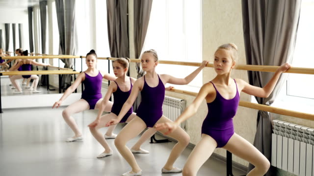 Small-girls-starting-ballet-dancers-in-leotards-are-doing-exercises-holding-ballet-bar-practicing-different-positions.-Dancing-class,-people-and-interior-concept.