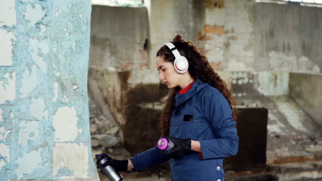 Kreative-junge-Frau-Graffiti-Maler-nutzt-Paint-Spray-um-zerstörten-Pfeiler-in-alten-leeren-Lagerhalle-zu-schmücken.-Mädchen-hört-Musik-über-Kopfhörer.