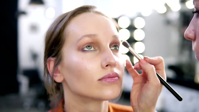 Close-up-of-a-model's-face-during-the-make-up-process.-Make-up-master-applying-light-brown-eyeshadows.-Blurred-view-of-white-lamps-on-background