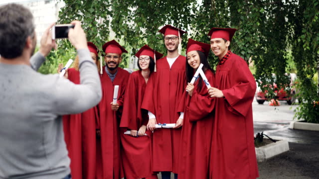 Proud-father-is-taking-pictures-of-graduating-students-with-smartphone-while-young-people-are-posing,-waving-hands-with-diplomas-and-gesturing.-Technology-and-education-concept.