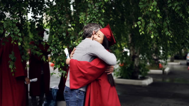 Loving-father-is-congratulating-his-daughter-on-graduation-day,-people-are-hugging-and-laughing-outdoors-while-other-graduates-are-visible-in-background.