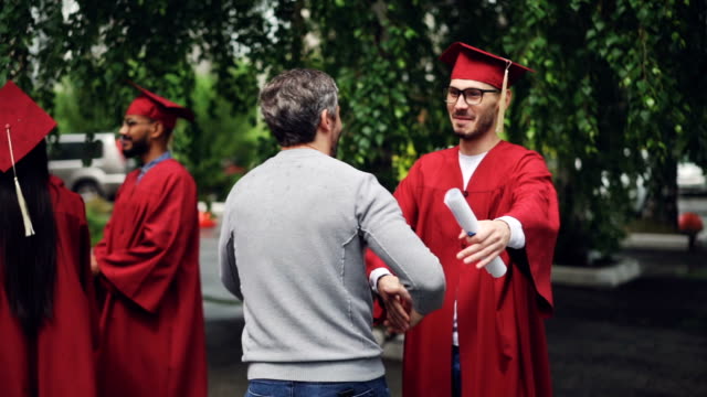 Lächelnde-Studium-Student-schüttelt-die-Hand-seines-Vaters-und-umarmt-ihn,-junger-Mann-in-Gläsern-trägt-Hut-und-Kleid-und-hält-Diplom.-Bildung-und-Erfolg-Konzept.