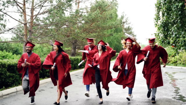 Slow-motion-of-emotional-graduates-happy-girls-and-guys-running-with-diplomas-and-laughing,-pretty-girl-is-taking-off-mortar-board-and-waving-it.-Small-rain-is-visible