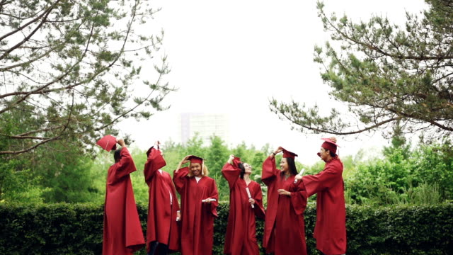 Slow-motion-of-happy-graduates-taking-off-mortarboard-caps,-throwing-in-the-air,-laughing-and-catching-them-then-jumping.-Education,-emotions-and-modern-youth-concept.