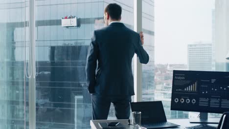 Following-Shot-of-the-Confident-Businessman-in-a-Suit-Walking-Through-His-Office-and-Looking-out-of-the-Window-Thoughtfully.-Stylish-Modern-Business-Office-with-Personal-Computer-and-Big-City-View.