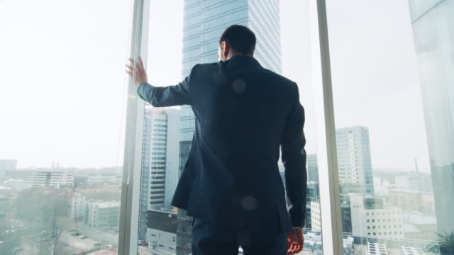 Thoughtful-Businessman-Wearing-Suit-Standing-in-His-Office,-Looking-out-of-the-Window-and-Contemplating-Next-Big-Business-Contract.-Major-City-Business-District-with-Panoramic-Window-View.-Blue-Colors.