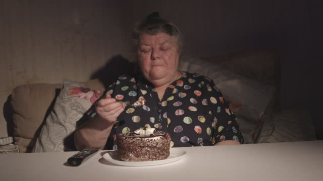 Senior-mujer-viendo-la-televisión-y-comiendo-pastel-en-un-cuarto-oscuro