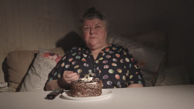Senior-mujer-viendo-la-televisión-y-comiendo-pastel-en-un-cuarto-oscuro