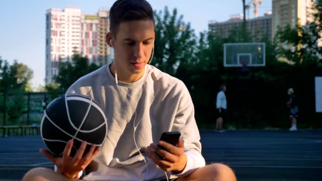 Handsome-young-man-sitting-on-basketball-court-and-typing-on-phone,-looking-at-camera,-holding-ball,-people-playing-in-background