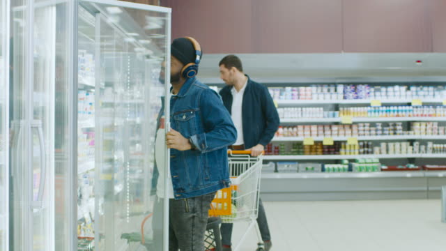 At-the-Supermarket:-Stylish-African-American-Guy-with-Shopping-Basket-Chooses-Products-in-the-Frozen-Goods-Section-of-the-Store.-He-Opens-Big-Glass-Door-Fridge.-Slow-Motion.