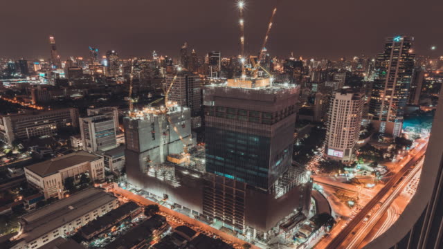 Day-to-night-holy-grail-time-lapse-of-building-construction-site-in-city,-traffic-light-trails-and-rain-at-the-end,-aerial-cityscape-view.-Construction-industry,-or-Asia-developing-country-concept
