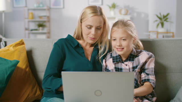 Beautiful-Young-Mom-and-Her-Cute-Little-Daugther-Use-Laptop-while-Sitting-on-a-Sofa-at-Home.-Family-Spending-Time-Together-Watching-Videos-and-Cartoons-on-Computer.