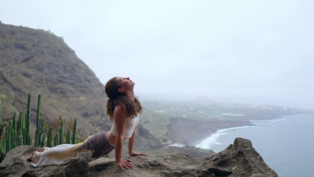 Mädchen-üben-von-Yoga-auf-den-Felsen-gegen-den-blauen-Himmel-und-das-azurblaue-Meer.-Frau-steht-auf-einem-Stein-in-einer-Brücke-Haltung.