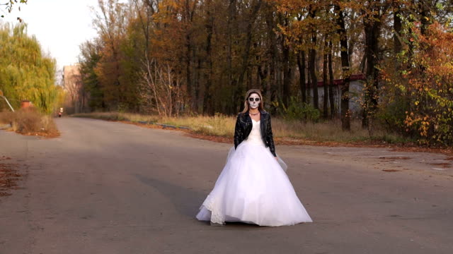A-girl-in-a-wedding-dress-with-a-skull-mask-on-an-empty-road.-Halloween.