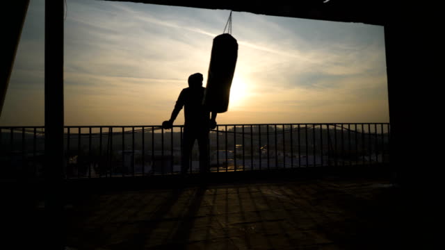 Silueta-de-bolsa-de-boxeo-mientras-que-hace-pivotar,-puesta-del-sol,-el-hombre-viene-y-mira-cerca,-deportista-practicando,-poder-formación,-chico-fuerte-duro-ejercicio,-ejercicios-de-fuerza,-entrenamiento,-mano,-soleado-día.