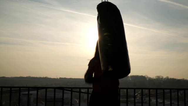 One-man-punches-hard-in-bag-on-boxing-training,-silhouette-of-boxer,-sun-shines,-sportsman-practicing,-power-training,-strong-guy-hard-exercising,-strength-exercises,-workout,-handheld,-sunny-day.