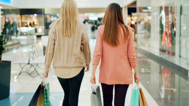 Rear-shot-of-slender-young-women-walking-in-shopping-mall-holding-paper-bags-and-looking-around-at-goods.-Happy-customers,-shiny-shop-windows-are-visible.