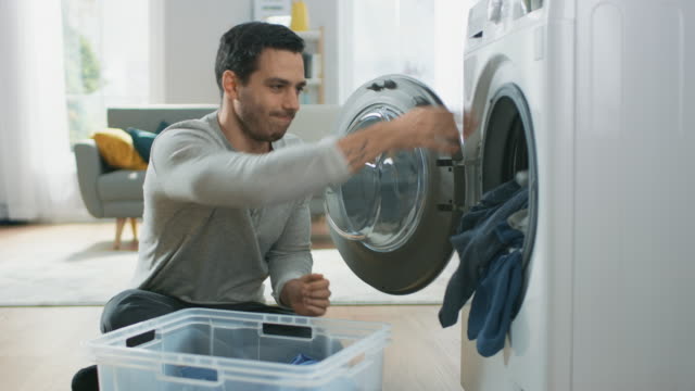 Handsome-Serious-Young-Man-in-Grey-Jeans-and-Coat-Sits-in-Front-of-a-Washing-Machine-at-Home.-He-Loads-the-Washer-with-Dirty-Laundry.-Bright-and-Spacious-Living-Room-with-Modern-Interior.