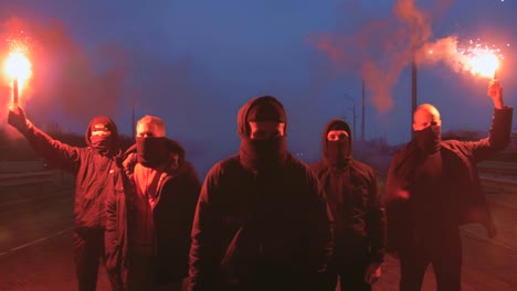 Gruppe-junger-Männer-in-Balaclavas-mit-rot-brennendem-Signal-flare-auf-der-Straße-unter-der-Brücke