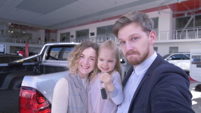 client-couple-with-cute-child-girl-with-keys-record-video-on-mobile-phone-near-new-purchased-car-in-auto-show