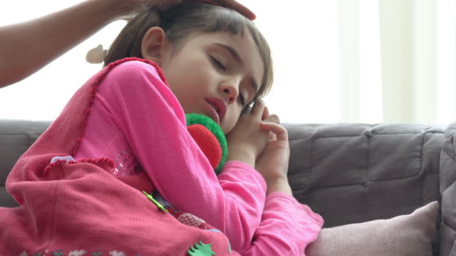 4K-Mittlere-Aufnahme-von-entzückenden-kleinen-Mädchen-krank-und-schlafen-auf-dem-Sofa-mit-Mutter-Hand-nehmen-Temperatur-auf-Tochter-Stirn.-Mutter-überprüft-Temperatur-der-kleinen-Tochter-Krankheit-mit-Fieber.