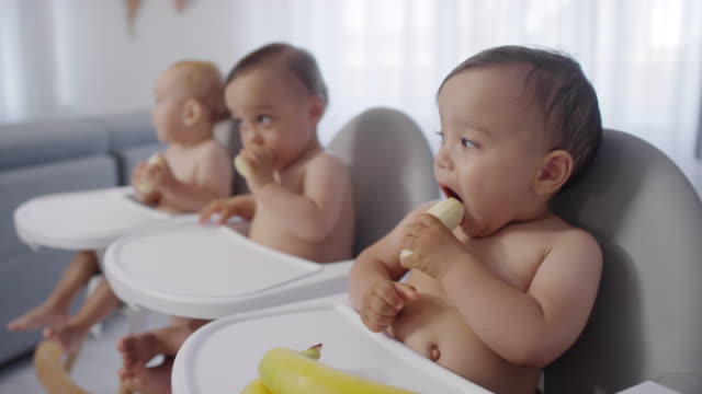 Gemischte-Rasse-Baby-Triplets-sitzen-in-Hochstühlen-und-Essen-Bananen