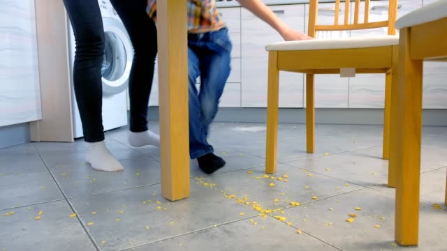 Mamá-regaña-a-su-hijo-por-comida-dispersa-en-el-piso-de-la-cocina-y-lo-hace-limpiar.-Limpie-las-escamas-de-maíz-del-suelo-juntas.