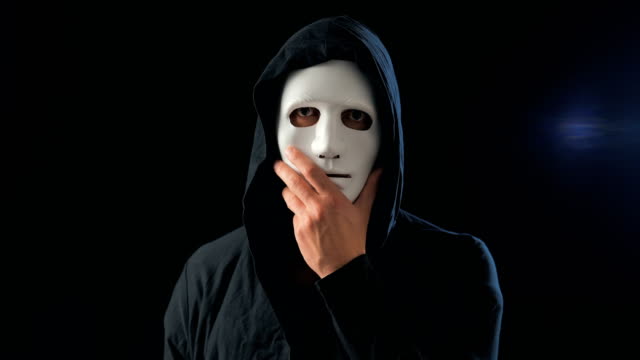 Hombre-anónimo-con-una-máscara-blanca-cubriendo-su-cara-y-una-capucha-negra-se-gira-y-mira-a-la-cámara.-Fondo-negro-oscuro.-Concepto-de-miembro-de-una-organización-secreta