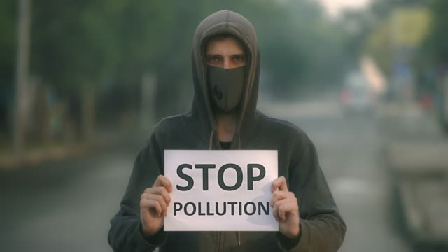 Man-in-respiratory-mask-look-at-camera-backdrop-road-with-banner-stop-pollution