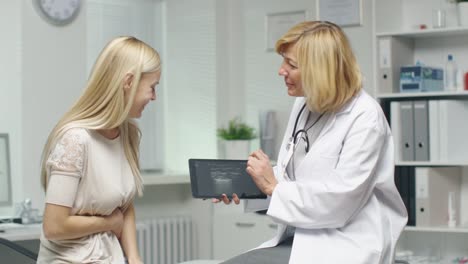 Mid-Adult-Female-Doctor-Consults-Young-Female-by-Showing-to-Her-Tablet-Computer-with-X-Ray.-Woman-Hodls-her-Stomach.-Both-Smile