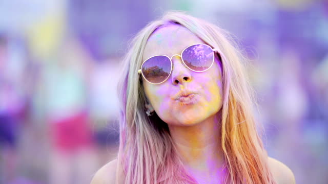 Mädchen-in-Sonnenbrille-bedeckt-in-bunte-Farbstoffe-Lächeln-auf-den-Lippen,-bläst-Luft-Kuss-an-Kamera