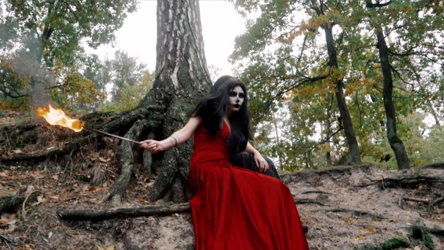 Zwei-Frauen-mit-scary-Halloween-Make-up-in-roten-und-schwarzen-Kleid-sitzt-in-der-Nähe-von-Baum-im-Waldpark-im-freien-Feuer-Fackel-in-der-hand-hält