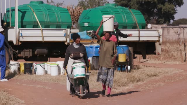 Two-african-woman-collecting-water-from-a--tanker-in-plastic-buckets-and-walking-back-to-their-homes