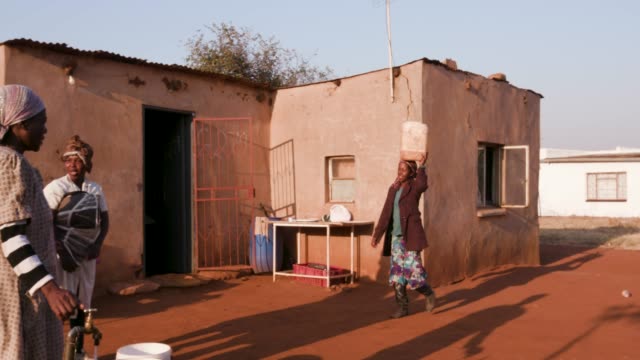 African-woman-arriving-at-a-house-to-collect-water-in-a-plastic-bucket,-while-others-stand-in-line-and-wait-their-turn