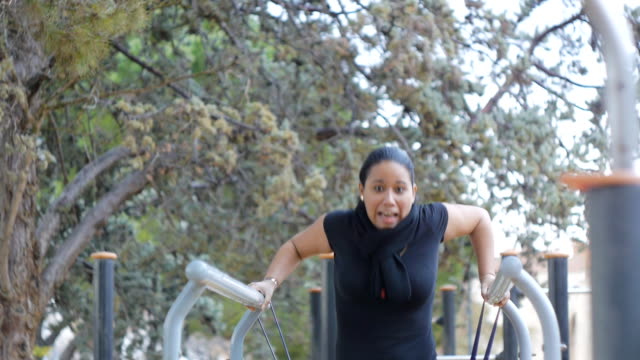 Street-workout-training.-Young-latin-american-woman-fails-falling-from-resistance-band-with-fright-trying-to-do-bar-dips-on-fitness-outdoor-gym-in-the-park.-The-girl-laughs-with-surprise.