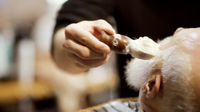 Barber-applies-shaving-cream-on-face-of-old-man-and-prepares-skin-to-shaving