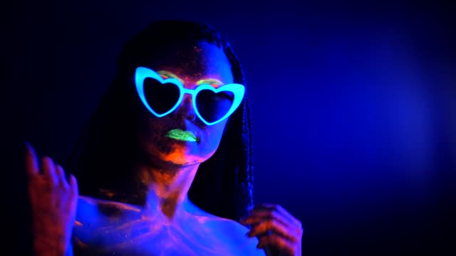 Fashion-sexy-dancer-with-heart-shaped-glasses-in-neon-light.-Fluorescent-makeup-glowing-under-ultraviolet-light.-Night-club,-party,-psychedelic-concepts.-Mysterious-woman-with-UV-painting
