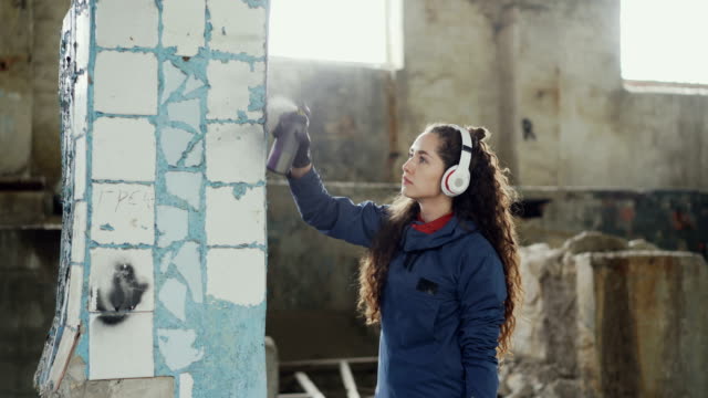 Attractive-girl-is-concentrated-on-decorating-old-dirty-column-with-graffiti-in-abandoned-building-using-aerosol-paint.-Young-woman-is-listening-to-music-with-headphones.
