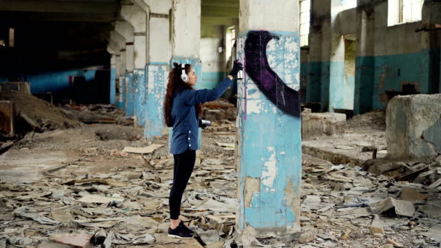 Urban-artist-is-decorating-column-in-abandoned-warehouse-with-abstract-image-using-aerosol-paint.-Girl-is-wearing-casual-clothing-and-listening-to-music-with-headphones.