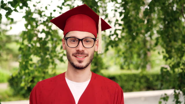 Portrait-of-bearded-young-man-graduating-student-wearing-glasses,-graduation-gown-and-mortar-board-smiling-and-looking-at-camera-standing-outdoors.-People-and-education-concept.