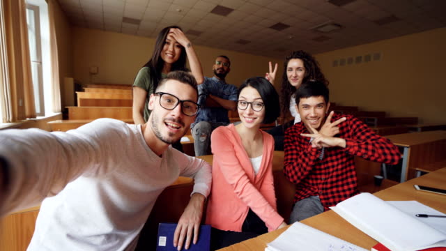 Point-of-view-shot-of-multiethnic-group-of-friends-taking-selfie-in-classroom-looking-at-camera,-posing-with-hand-gestures-and-smiling.-Modern-technology-and-millennials-concept.