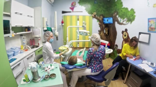 Child-with-a-mother-at-a-dentist's-reception.-The-girl-lies-in-the-chair,-behind-her-mother.-The-doctor-works-with-an-assistant.-Procedure-for-drilling-a-tooth.-Setting-a-temporary-seal.